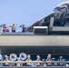 The image shows a Marvel-themed event, likely at a Disney theme park or cruise ship, featuring costumed characters including Captain America on an upper deck alongside what appears to be a military band member and another character. A large Avengers-branded aircraft or quinjet prop is prominently displayed on the upper level. Below, a marching band in white uniforms performs with brass instruments including tubas and trumpets.