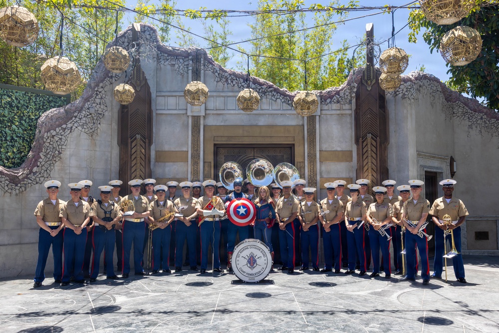 A U.S. Marine Corps band from Camp Pendleton, California poses for a group photo in front of an ornate themed structure, likely at a Disney theme park (Avengers Campus area). The group includes approximately 25-30 uniformed Marines holding various instruments, flanked by two costumed characters including Captain America. A large bass drum in the foreground bears the inscription referencing the 1st Marine Division Band, Camp Pendleton, CA.