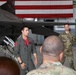 A military briefing is taking place inside an aircraft hangar where a pilot in an olive flight suit is addressing a group of personnel. The setting features an F-16 Fighting Falcon fighter jet and a large American flag displayed on the hangar wall in the background. Multiple service members in OCP (Operational Camouflage Pattern) uniforms are present as the audience.