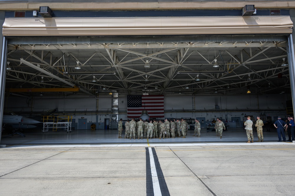 A large military aircraft hangar with its bay doors fully open, revealing a group of uniformed personnel gathered inside in front of a military jet aircraft. An American flag is prominently displayed on the back wall of the hangar. The scene appears to depict a formal or ceremonial military gathering at a U.S. Air Force installation.