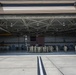A large military aircraft hangar with its bay doors fully open, revealing a group of uniformed personnel gathered inside in front of a military jet aircraft. An American flag is prominently displayed on the back wall of the hangar. The scene appears to depict a formal or ceremonial military gathering at a U.S. Air Force installation.