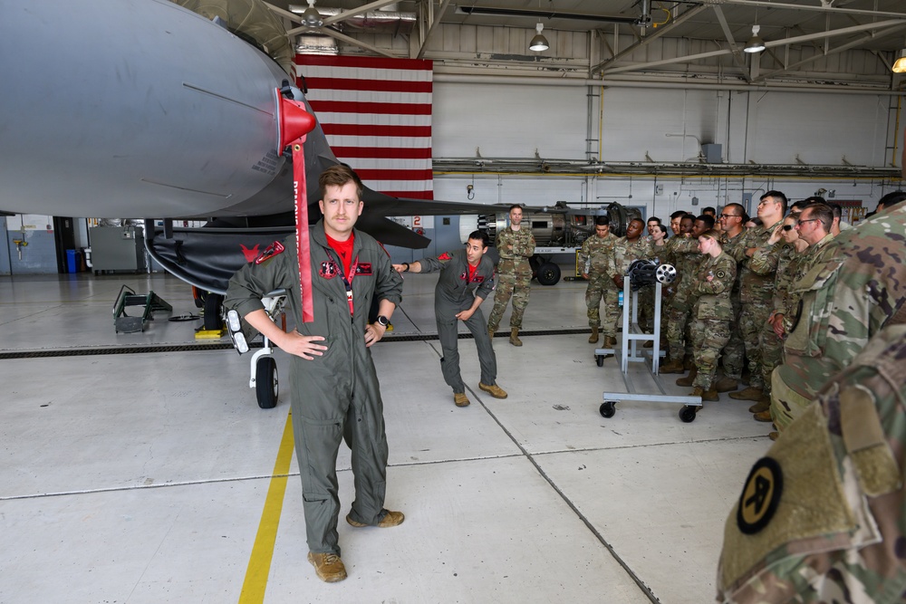 A military aviation briefing or tour is taking place inside an aircraft hangar. A pilot or crew member in an olive flight suit with a red shirt underneath stands near the nose of a gray military jet, appearing to address a large group of soldiers in camouflage uniforms. An American flag is visible in the background, and the hangar is well-lit with overhead lighting.