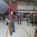 A military aviation briefing or tour is taking place inside an aircraft hangar. A pilot or crew member in an olive flight suit with a red shirt underneath stands near the nose of a gray military jet, appearing to address a large group of soldiers in camouflage uniforms. An American flag is visible in the background, and the hangar is well-lit with overhead lighting.