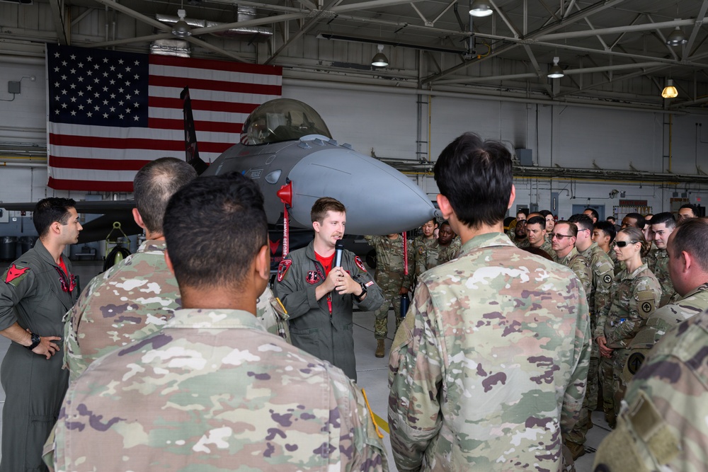 A military briefing or presentation is taking place inside an aircraft hangar, where a person in a flight suit is speaking into a microphone to a large group of uniformed personnel in OCP (Operational Camouflage Pattern) uniforms. A gray fighter jet, consistent with an F-16 Fighting Falcon, is prominently displayed in the background alongside a large American flag. The setting suggests a formal orientation, tour, or unit briefing event.