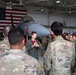 A military briefing or presentation is taking place inside an aircraft hangar, where a person in a flight suit is speaking into a microphone to a large group of uniformed personnel in OCP (Operational Camouflage Pattern) uniforms. A gray fighter jet, consistent with an F-16 Fighting Falcon, is prominently displayed in the background alongside a large American flag. The setting suggests a formal orientation, tour, or unit briefing event.