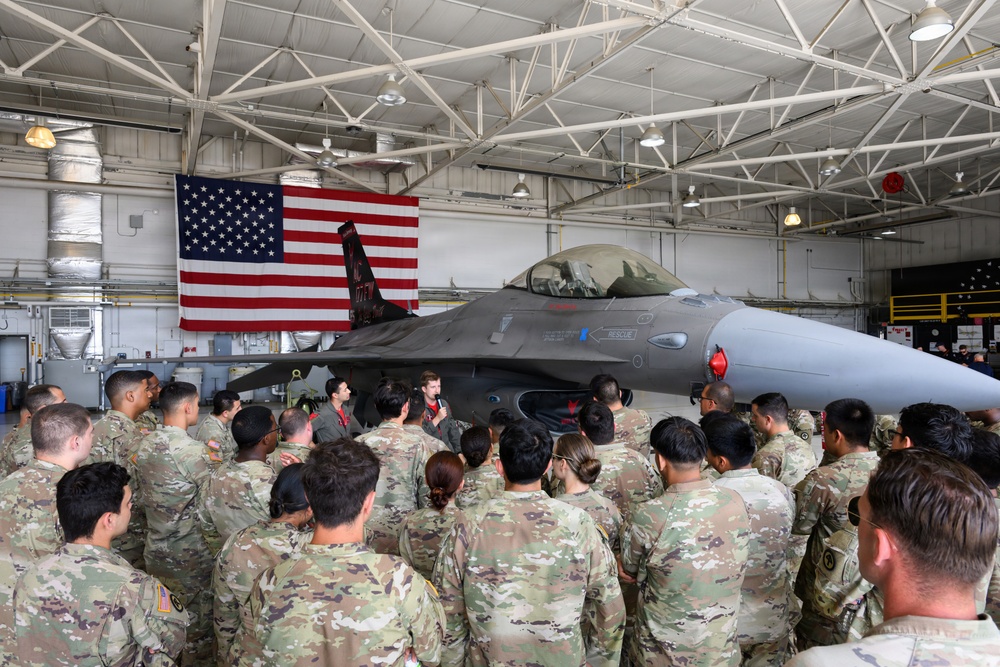 A large group of U.S. military personnel in MultiCam/OCP camouflage uniforms are gathered inside an aircraft hangar, listening to a briefing by two individuals in flight suits standing in front of an F-16 Fighting Falcon jet. A large American flag is displayed prominently on the hangar wall behind the aircraft. The setting appears to be an official military orientation or educational event at an Air Force installation.