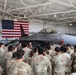 A large group of U.S. military personnel in MultiCam/OCP camouflage uniforms are gathered inside an aircraft hangar, listening to a briefing by two individuals in flight suits standing in front of an F-16 Fighting Falcon jet. A large American flag is displayed prominently on the hangar wall behind the aircraft. The setting appears to be an official military orientation or educational event at an Air Force installation.