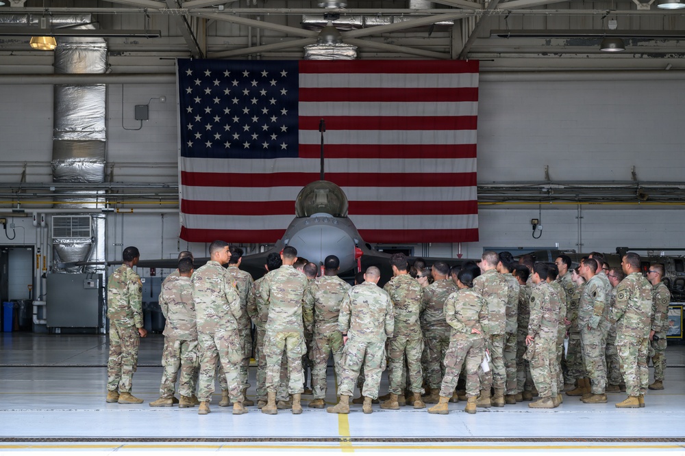 A large group of military personnel in OCP (Operational Camouflage Pattern) uniforms are gathered inside an aircraft hangar, facing away from the camera toward a military jet aircraft. A large American flag is prominently displayed on the hangar wall behind the aircraft. The setting appears to be a formal or ceremonial gathering at a U.S. military air base.