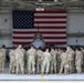 A large group of military personnel in OCP (Operational Camouflage Pattern) uniforms are gathered inside an aircraft hangar, facing away from the camera toward a military jet aircraft. A large American flag is prominently displayed on the hangar wall behind the aircraft. The setting appears to be a formal or ceremonial gathering at a U.S. military air base.