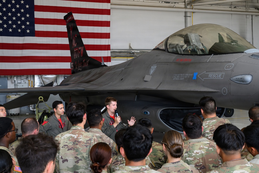 A military briefing or presentation is taking place inside an aircraft hangar, with a pilot speaking into a microphone in front of an F-16 Fighting Falcon jet. A large American flag is displayed prominently on the hangar wall behind the aircraft. A crowd of uniformed military personnel in camouflage (OCP/MultiCam) uniforms are gathered to listen, along with two individuals in flight suits.