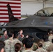 A military briefing or presentation is taking place inside an aircraft hangar, with a pilot speaking into a microphone in front of an F-16 Fighting Falcon jet. A large American flag is displayed prominently on the hangar wall behind the aircraft. A crowd of uniformed military personnel in camouflage (OCP/MultiCam) uniforms are gathered to listen, along with two individuals in flight suits.