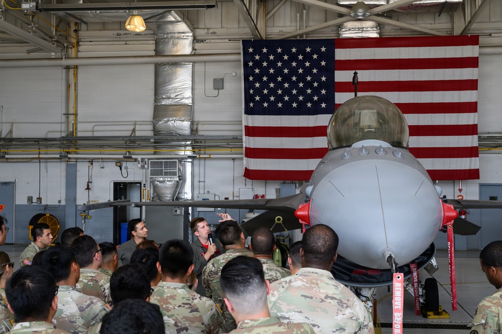 A military briefing is taking place inside an aircraft hangar where a speaker holding a microphone addresses a large group of uniformed personnel in camouflage (OCP/MultiCam) pattern uniforms. The briefing is conducted in front of what appears to be an F-16 Fighting Falcon fighter jet, with a large American flag displayed prominently on the hangar wall behind it. Red safety streamers and 'Remove Before Flight' tags are visible on the aircraft.
