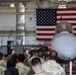 A military briefing is taking place inside an aircraft hangar where a speaker holding a microphone addresses a large group of uniformed personnel in camouflage (OCP/MultiCam) pattern uniforms. The briefing is conducted in front of what appears to be an F-16 Fighting Falcon fighter jet, with a large American flag displayed prominently on the hangar wall behind it. Red safety streamers and 'Remove Before Flight' tags are visible on the aircraft.
