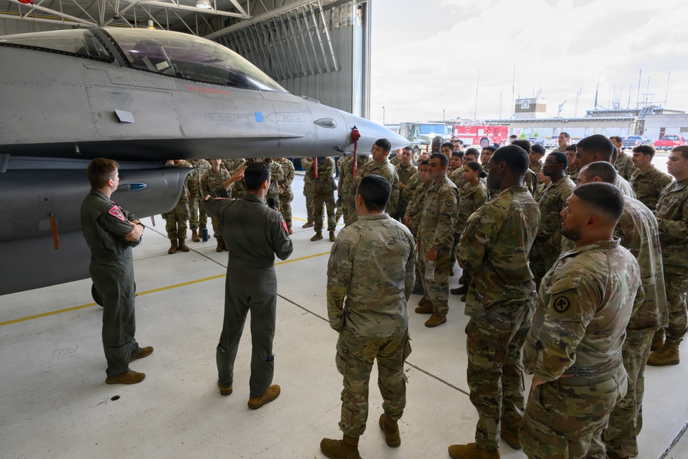 Two pilots in olive-green flight suits are conducting a briefing or tour for a large group of soldiers in OCP (Operational Camouflage Pattern) uniforms inside an aircraft hangar. The focal point is an F-16 Fighting Falcon fighter jet parked in the hangar. This appears to be an educational or orientation event where ground troops are being introduced to Air Force aircraft and capabilities.