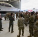 Two pilots in olive-green flight suits are conducting a briefing or tour for a large group of soldiers in OCP (Operational Camouflage Pattern) uniforms inside an aircraft hangar. The focal point is an F-16 Fighting Falcon fighter jet parked in the hangar. This appears to be an educational or orientation event where ground troops are being introduced to Air Force aircraft and capabilities.