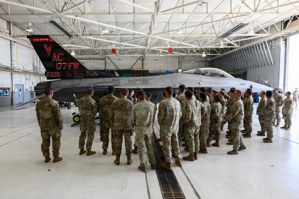 A large group of military personnel in camouflage uniforms are gathered in an aircraft hangar around an F-16 Fighting Falcon jet. The aircraft bears the markings of the 177th Fighter Wing (177 FW) from New Jersey Air National Guard, nicknamed 'The Jersey Devils,' with tail code AC and serial number AF 87-225. A briefing or orientation appears to be taking place in front of the aircraft.