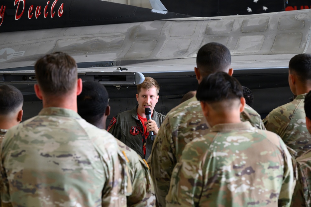 A military aviator in a flight suit is addressing a group of soldiers in OCP (Operational Camouflage Pattern) uniforms inside an aircraft hangar. He is holding a microphone and speaking in front of what appears to be a military jet aircraft with 'Devils' visible on its fuselage. The setting suggests a briefing, orientation, or demonstration event.