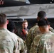 A military aviator in a flight suit is addressing a group of soldiers in OCP (Operational Camouflage Pattern) uniforms inside an aircraft hangar. He is holding a microphone and speaking in front of what appears to be a military jet aircraft with 'Devils' visible on its fuselage. The setting suggests a briefing, orientation, or demonstration event.