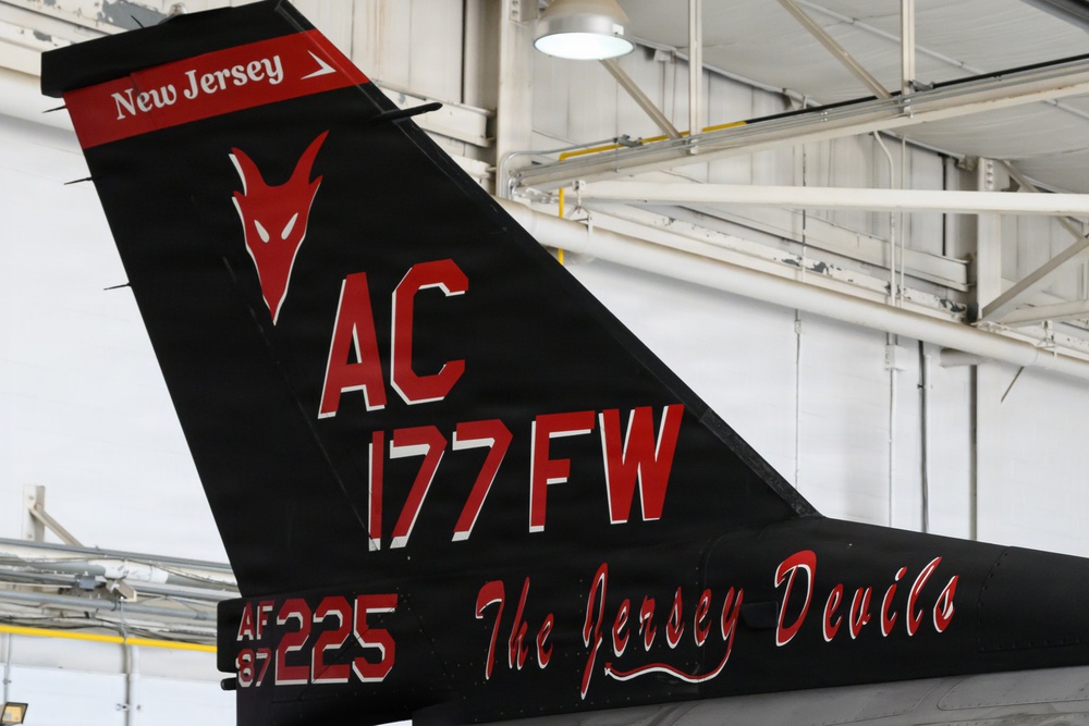 The image shows the tail section of a military jet aircraft, specifically belonging to the 177th Fighter Wing of the New Jersey Air National Guard. The tail features a distinctive black paint scheme with red markings, including the unit's 'Jersey Devils' nickname and a stylized devil mascot. The aircraft is photographed inside a hangar.