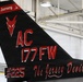 The image shows the tail section of a military jet aircraft, specifically belonging to the 177th Fighter Wing of the New Jersey Air National Guard. The tail features a distinctive black paint scheme with red markings, including the unit's 'Jersey Devils' nickname and a stylized devil mascot. The aircraft is photographed inside a hangar.