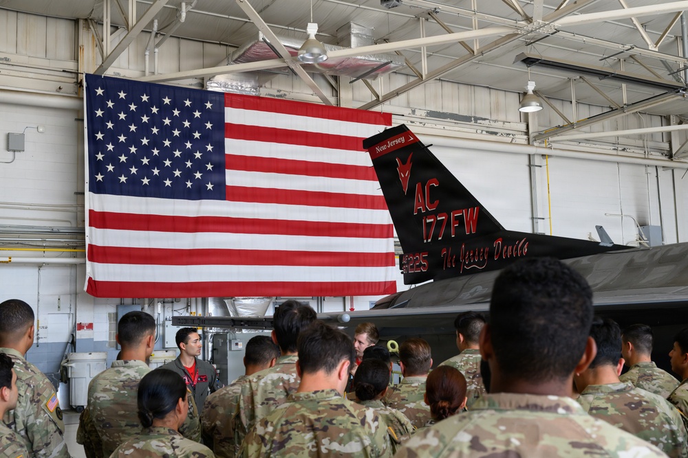 A group of military personnel in camouflage uniforms are gathered inside an aircraft hangar, facing a speaker in a flight suit. Behind them is a large American flag and a black-painted fighter jet tail section bearing the markings of the 177th Fighter Wing, New Jersey Air National Guard, nicknamed 'The Jersey Devils'. The setting appears to be a formal briefing or orientation event.