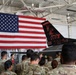 A group of military personnel in camouflage uniforms are gathered inside an aircraft hangar, facing a speaker in a flight suit. Behind them is a large American flag and a black-painted fighter jet tail section bearing the markings of the 177th Fighter Wing, New Jersey Air National Guard, nicknamed 'The Jersey Devils'. The setting appears to be a formal briefing or orientation event.
