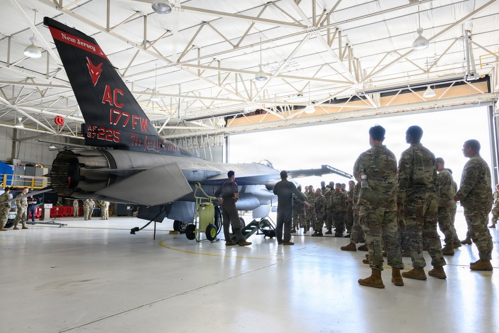 Military personnel in camouflage uniforms are gathered in an aircraft hangar observing a specially painted F-16 Fighting Jet belonging to the 177th Fighter Wing of the New Jersey Air National Guard. The aircraft features a distinctive black and red tail design with 'AC 177FW' markings, the serial number AF 87-225, and the text 'The Jersey Devil' along with a devil logo. The scene appears to be a formal presentation or briefing event.