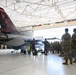 Military personnel in camouflage uniforms are gathered in an aircraft hangar observing a specially painted F-16 Fighting Jet belonging to the 177th Fighter Wing of the New Jersey Air National Guard. The aircraft features a distinctive black and red tail design with 'AC 177FW' markings, the serial number AF 87-225, and the text 'The Jersey Devil' along with a devil logo. The scene appears to be a formal presentation or briefing event.