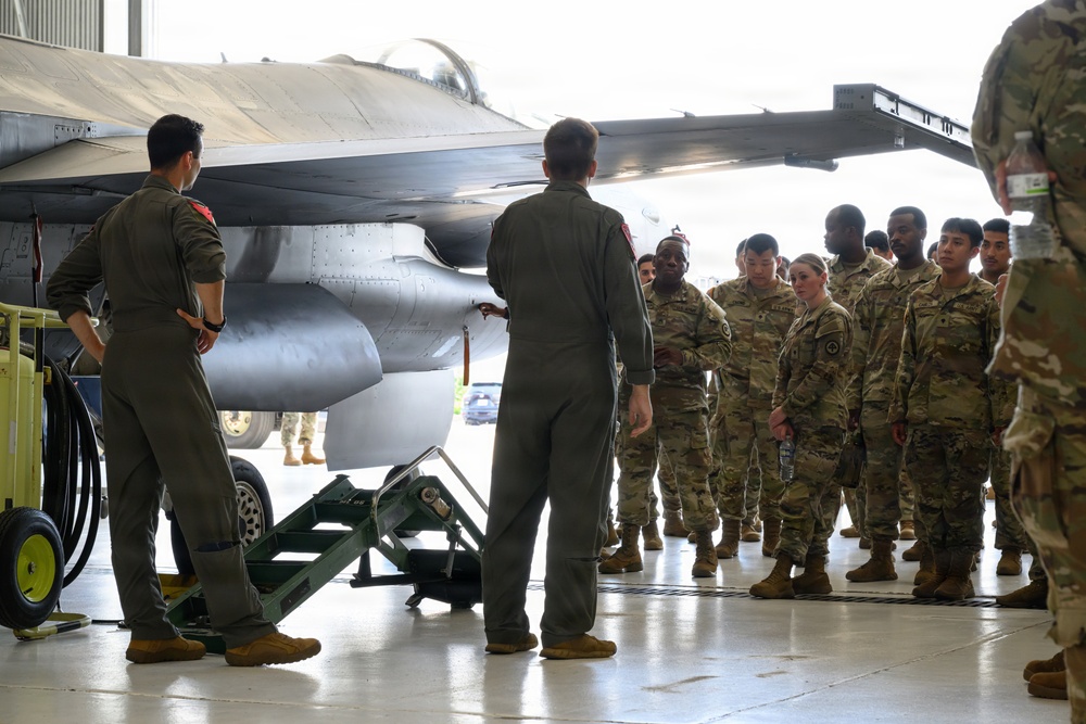 Two pilots in olive-green flight suits are conducting a briefing or walkthrough of an F-16 Fighting Falcon aircraft inside a military hangar. A group of approximately 15-20 soldiers in MultiCam/OCP camouflage uniforms attentively observe the presentation. This appears to be an orientation, familiarization training, or cross-branch educational event.