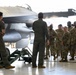 Two pilots in olive-green flight suits are conducting a briefing or walkthrough of an F-16 Fighting Falcon aircraft inside a military hangar. A group of approximately 15-20 soldiers in MultiCam/OCP camouflage uniforms attentively observe the presentation. This appears to be an orientation, familiarization training, or cross-branch educational event.