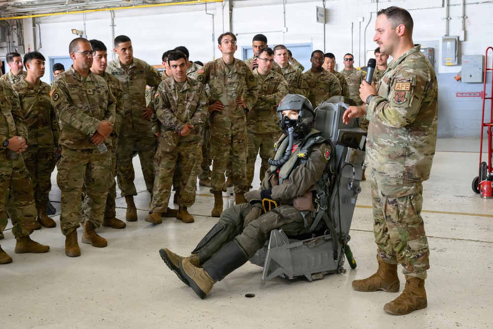 A military instructor wearing an 'AFE' (Aircrew Flight Equipment) patch is conducting a briefing or demonstration for a group of soldiers in a hangar facility. A mannequin or person is seated in what appears to be an ejection seat, wearing full flight gear including a helmet, oxygen mask, and flight suit. The audience consists of approximately 20 uniformed military personnel in OCP (Operational Camouflage Pattern) uniforms attentively observing the presentation.