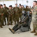 A military instructor wearing an 'AFE' (Aircrew Flight Equipment) patch is conducting a briefing or demonstration for a group of soldiers in a hangar facility. A mannequin or person is seated in what appears to be an ejection seat, wearing full flight gear including a helmet, oxygen mask, and flight suit. The audience consists of approximately 20 uniformed military personnel in OCP (Operational Camouflage Pattern) uniforms attentively observing the presentation.