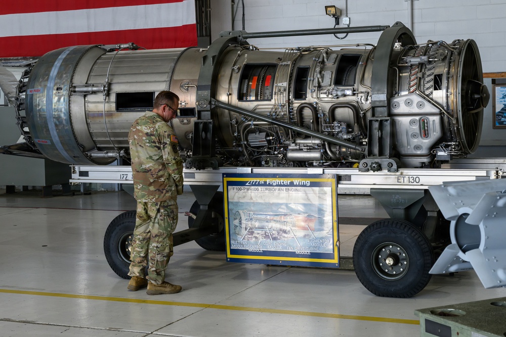 A military service member in camouflage uniform examines an F100-PW-100 turbofan jet engine mounted on a wheeled transport cart inside a hangar. The engine is displayed in a cutaway/cross-section configuration, exposing its internal components. A placard on the cart identifies it as belonging to a Fighter Wing and labels the engine's major modules.