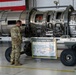 A military service member in camouflage uniform examines an F100-PW-100 turbofan jet engine mounted on a wheeled transport cart inside a hangar. The engine is displayed in a cutaway/cross-section configuration, exposing its internal components. A placard on the cart identifies it as belonging to a Fighter Wing and labels the engine's major modules.
