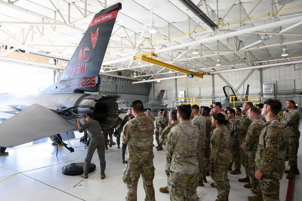 A group of military personnel in camouflage uniforms are gathered inside an aircraft hangar for what appears to be a briefing or demonstration around an F-16 Fighting Falcon jet. The aircraft's tail markings clearly identify it as belonging to the 177th Fighter Wing (177 FW) of the New Jersey Air National Guard, with tail code 'AC' and number 225. One individual in a flight suit appears to be conducting a hands-on demonstration or inspection of the aircraft's underside.