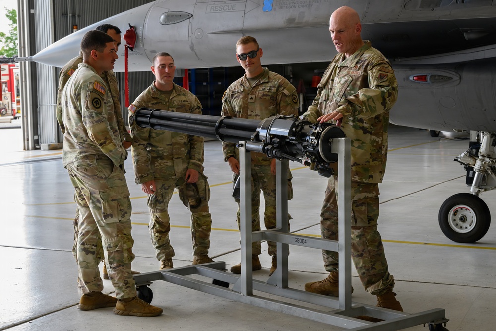 Five military personnel in OCP (Operational Camouflage Pattern) uniforms are gathered in an aircraft hangar examining a multi-barrel rotary cannon mounted on a wheeled maintenance stand. One individual, appearing to be a senior non-commissioned officer or officer, is gesturing toward the weapon system while briefing the others. A military jet aircraft, consistent with an F-16 Fighting Falcon, is visible in the background.