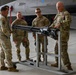 Five military personnel in OCP (Operational Camouflage Pattern) uniforms are gathered in an aircraft hangar examining a multi-barrel rotary cannon mounted on a wheeled maintenance stand. One individual, appearing to be a senior non-commissioned officer or officer, is gesturing toward the weapon system while briefing the others. A military jet aircraft, consistent with an F-16 Fighting Falcon, is visible in the background.