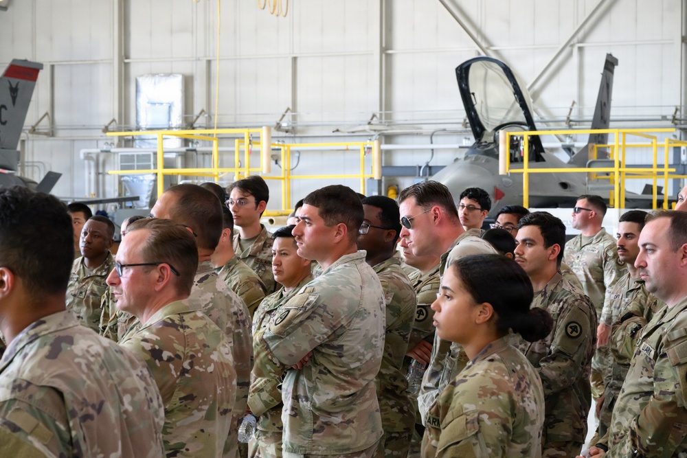 A large group of military personnel in U.S. Army OCP (Operational Camouflage Pattern) uniforms are gathered inside a military aircraft hangar. They appear to be attending a briefing or formation event, standing attentively and facing a common direction. In the background, at least one military jet aircraft (appearing to be an F-16 or similar fighter jet) is visible on the hangar floor.