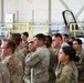 A large group of military personnel in U.S. Army OCP (Operational Camouflage Pattern) uniforms are gathered inside a military aircraft hangar. They appear to be attending a briefing or formation event, standing attentively and facing a common direction. In the background, at least one military jet aircraft (appearing to be an F-16 or similar fighter jet) is visible on the hangar floor.