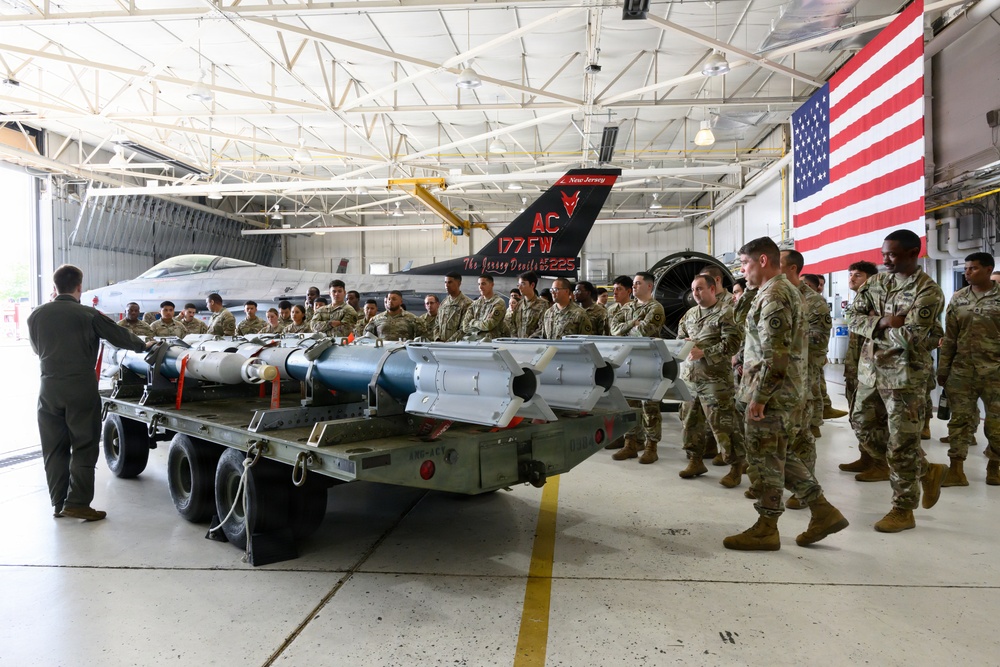 A military briefing or demonstration is taking place inside an aircraft hangar at what appears to be the 177th Fighter Wing (New Jersey Air National Guard), known as 'The Jersey Devils.' A group of uniformed service members in camouflage (OCP) uniforms are gathered around a weapons cart loaded with air-to-air and air-to-ground munitions, being addressed by an instructor in a flight suit. An F-16 Fighting Falcon aircraft and a large American flag are visible in the background.