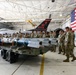 A military briefing or demonstration is taking place inside an aircraft hangar at what appears to be the 177th Fighter Wing (New Jersey Air National Guard), known as 'The Jersey Devils.' A group of uniformed service members in camouflage (OCP) uniforms are gathered around a weapons cart loaded with air-to-air and air-to-ground munitions, being addressed by an instructor in a flight suit. An F-16 Fighting Falcon aircraft and a large American flag are visible in the background.