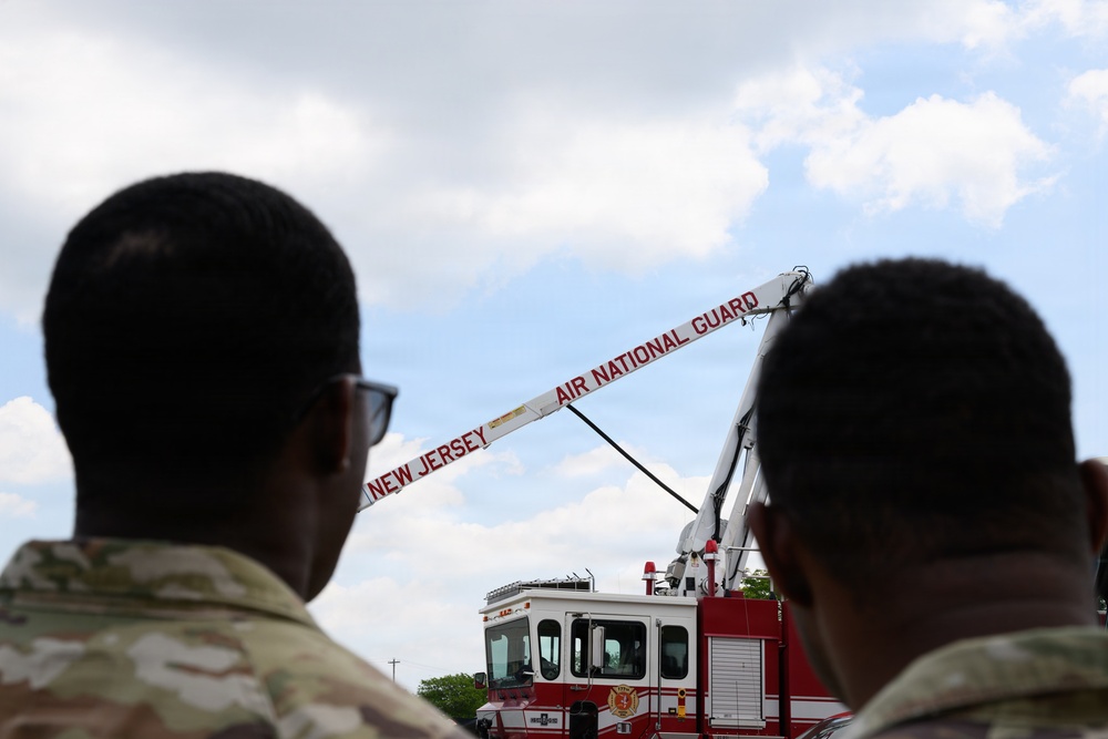 Two military personnel in camouflage uniforms observe a New Jersey Air National Guard fire truck with its aerial ladder extended. The fire truck is red and white, prominently labeled 'NEW JERSEY AIR NATIONAL GUARD' along the ladder boom. The scene appears to be an outdoor demonstration or equipment display under a partly cloudy sky.