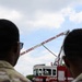 Two military personnel in camouflage uniforms observe a New Jersey Air National Guard fire truck with its aerial ladder extended. The fire truck is red and white, prominently labeled 'NEW JERSEY AIR NATIONAL GUARD' along the ladder boom. The scene appears to be an outdoor demonstration or equipment display under a partly cloudy sky.