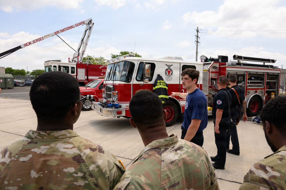 A group of military personnel in camouflage uniforms are receiving a briefing or tour from firefighters in front of New Jersey Air National Guard fire trucks. The scene appears to be a training or familiarization exercise at a military installation. A young man in a blue t-shirt is gesturing and speaking to the group near the fire apparatus.