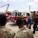 A group of military personnel in camouflage uniforms are receiving a briefing or tour from firefighters in front of New Jersey Air National Guard fire trucks. The scene appears to be a training or familiarization exercise at a military installation. A young man in a blue t-shirt is gesturing and speaking to the group near the fire apparatus.