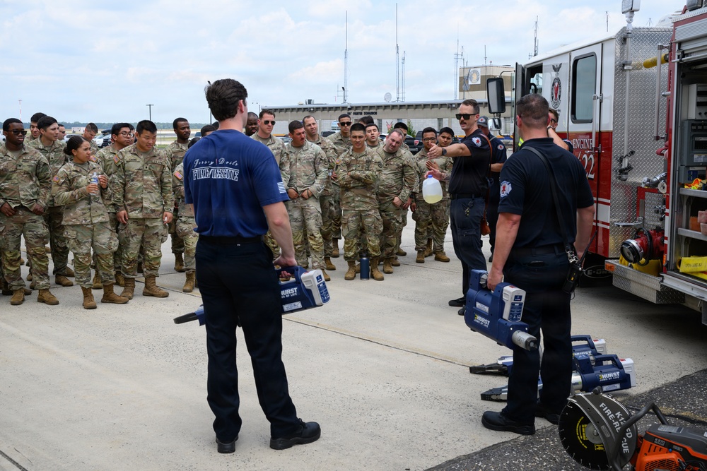 A group of military personnel in camouflage uniforms are receiving a demonstration or briefing from firefighters wearing dark navy blue uniforms marked 'Fire/Rescue'. The firefighters are holding and displaying Hurst rescue tools (commonly known as 'Jaws of Life') next to a red fire engine. This appears to be a training or familiarization event conducted outdoors on a paved area, likely at a military installation.