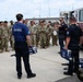 A group of military personnel in camouflage uniforms are receiving a demonstration or briefing from firefighters wearing dark navy blue uniforms marked 'Fire/Rescue'. The firefighters are holding and displaying Hurst rescue tools (commonly known as 'Jaws of Life') next to a red fire engine. This appears to be a training or familiarization event conducted outdoors on a paved area, likely at a military installation.