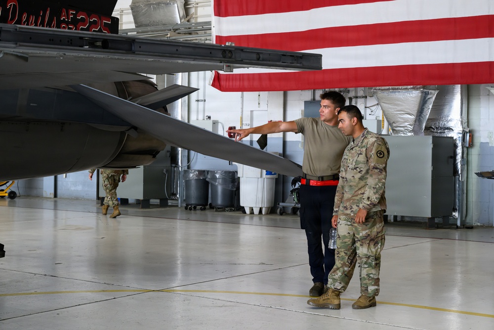 Two military personnel are standing in an aircraft hangar examining the underside of a military jet aircraft, with one individual pointing toward a component on the aircraft. A large American flag is draped in the background of the hangar. A third uniformed individual is visible in the background walking near the aircraft.