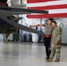 Two military personnel are standing in an aircraft hangar examining the underside of a military jet aircraft, with one individual pointing toward a component on the aircraft. A large American flag is draped in the background of the hangar. A third uniformed individual is visible in the background walking near the aircraft.