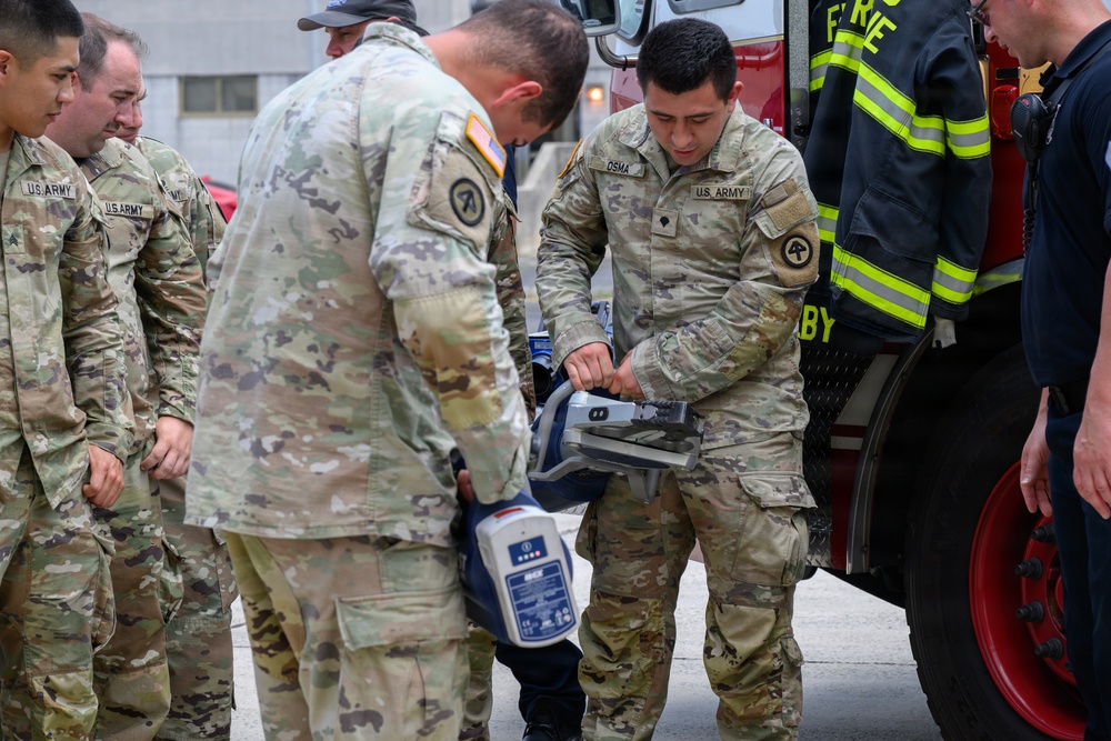 Several U.S. Army soldiers in OCP (Operational Camouflage Pattern) uniforms are gathered near a fire truck, examining or handling a medical/electronic device. One soldier, identified by name tape as 'OSMA,' is actively handling what appears to be a portable medical monitor or defibrillator. A firefighter in high-visibility gear is visible on the right side of the frame, suggesting an interagency training or coordination exercise.